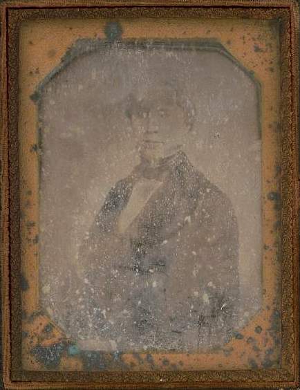 A daguerreotype of a well-dressed man, but it has been eaten at so severely by wear and age that it is difficult to see his form and his face.